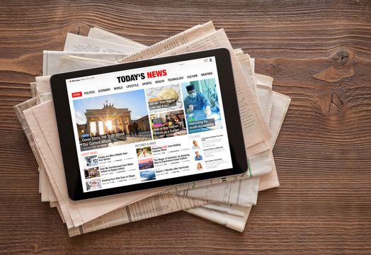 Tablet showing the day's news on top of a stack of newspapers